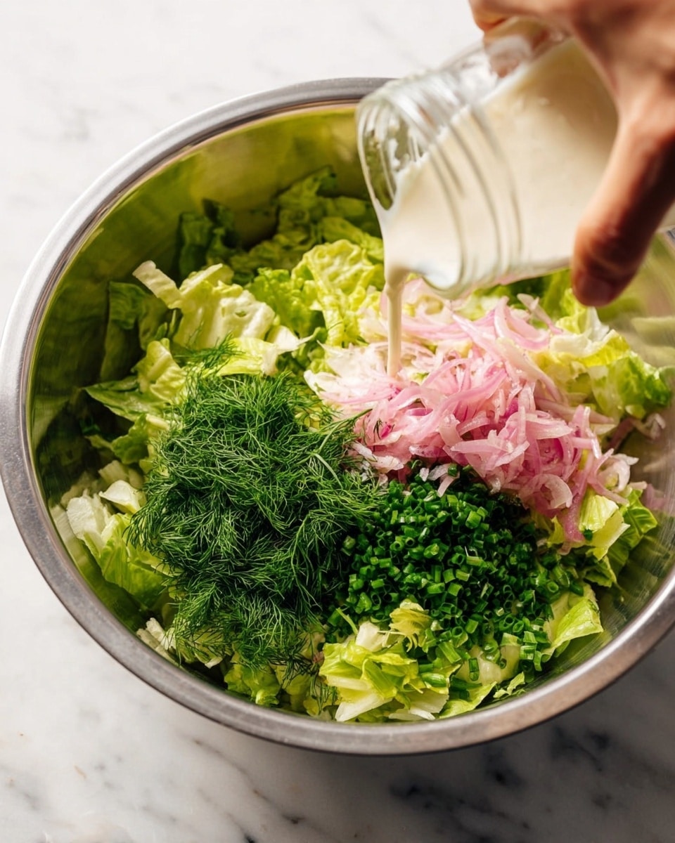 A metal mixing bowl sits on a white marbled surface filled with layers of fresh green lettuce pieces as the base. On top of the lettuce, there are three separate piles: bright green chopped chives, finely chopped dark green dill, and a small heap of pale pink pickled onion slices. A woman's hand is shown pouring a clear liquid from a glass jar over the ingredients, ready to mix them all together. The metal bowl reflects some light, adding a shiny texture to the scene. photo taken with an iphone --ar 4:5 --v 7