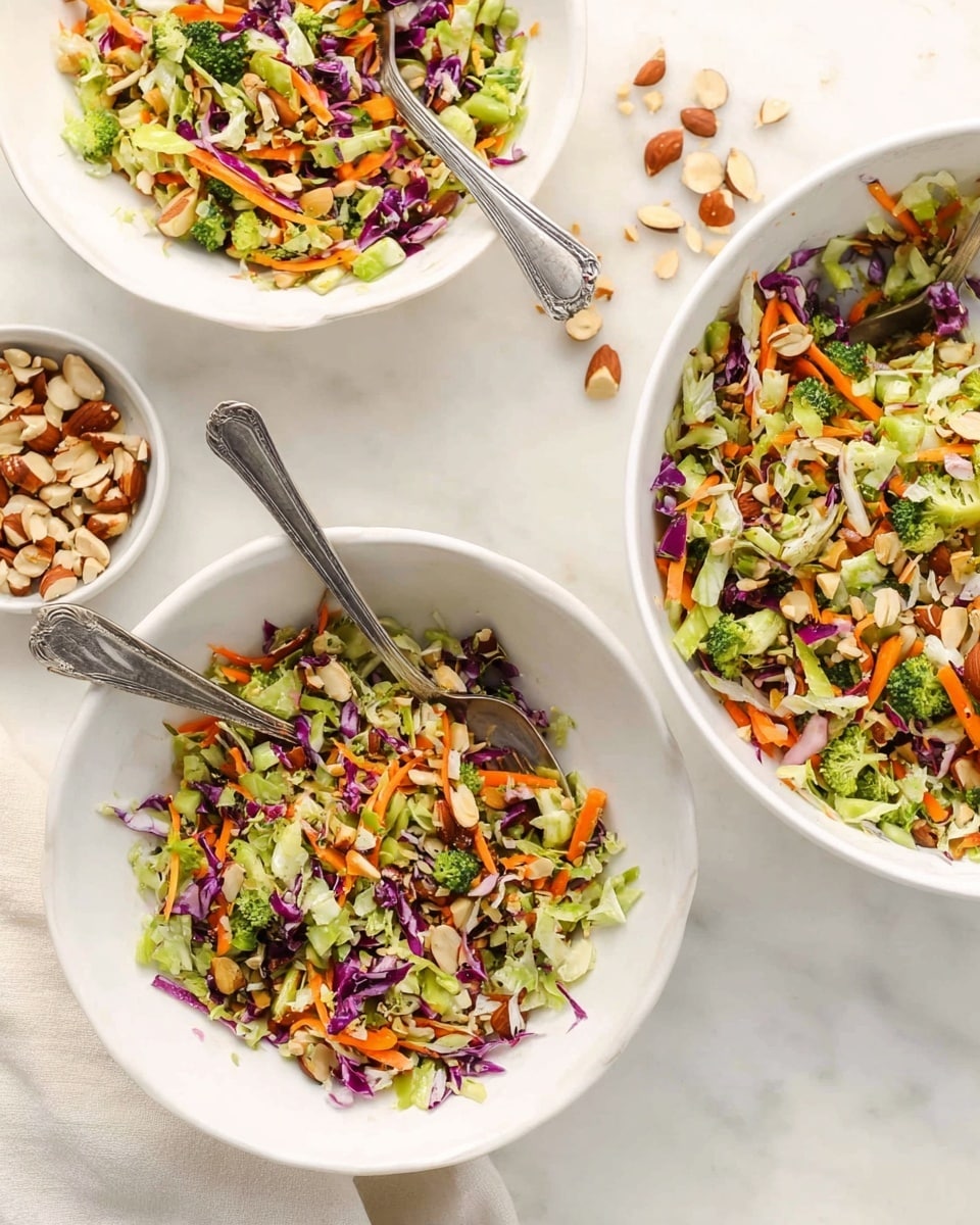 The image shows a colorful salad served in three white bowls on a white marbled surface. Each bowl contains a vibrant mix of finely chopped vegetables and nuts, including orange carrot shreds, green celery, broccoli pieces, purple cabbage, and brown almond pieces, all mixed evenly. Two silver forks rest inside two of the bowls, angled outward. To the side, there is a small white bowl containing extra chopped almonds. The overall look is fresh and bright, with the salad layers showing varied textures from crunchy nuts to crisp vegetables. Photo taken with an iphone --ar 4:5 --v 7