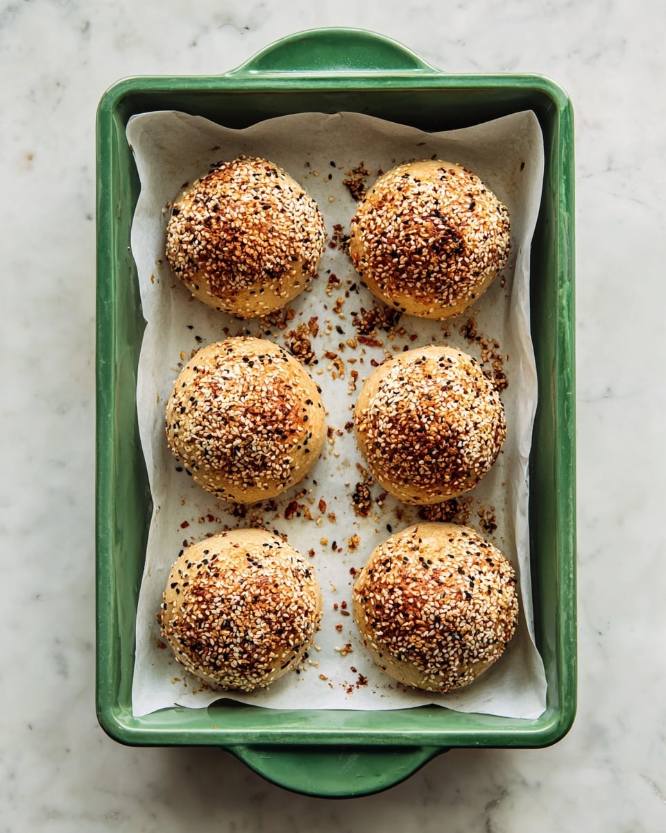 The image shows a green baking tray with a white liner inside, holding eight round baked buns arranged in two neat columns of four. Each bun is golden brown and covered with a mix of toasted sesame seeds, white sesame seeds, and other small seeds, giving a textured and crunchy look. The buns have an uneven, slightly rough surface with some visible cracks and crumbs scattered around them. The tray is set on a white marbled surface, creating a clean and bright background. Photo taken with an iphone --ar 4:5 --v 7