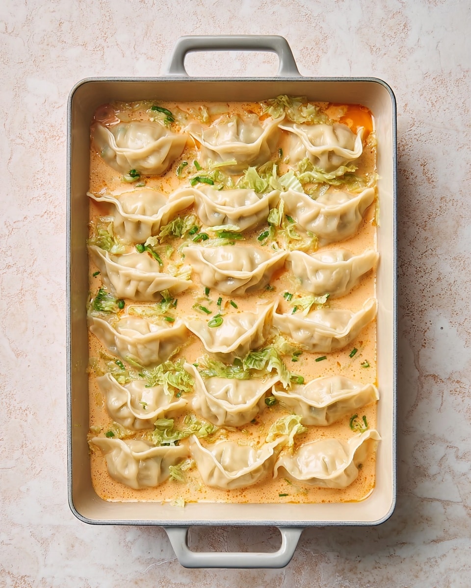 A light gray rectangular baking dish with two handles holds twenty dumplings arranged in neat rows on top of a creamy light orange sauce with finely chopped green cabbage pieces scattered throughout. The dumplings have pale, smooth wrappers with crimped edges and slight folds visible, floating gently in the sauce. The background is a white marbled surface with a soft, warm light enhancing the pastel colors in the scene. Photo taken with an iphone --ar 4:5 --v 7