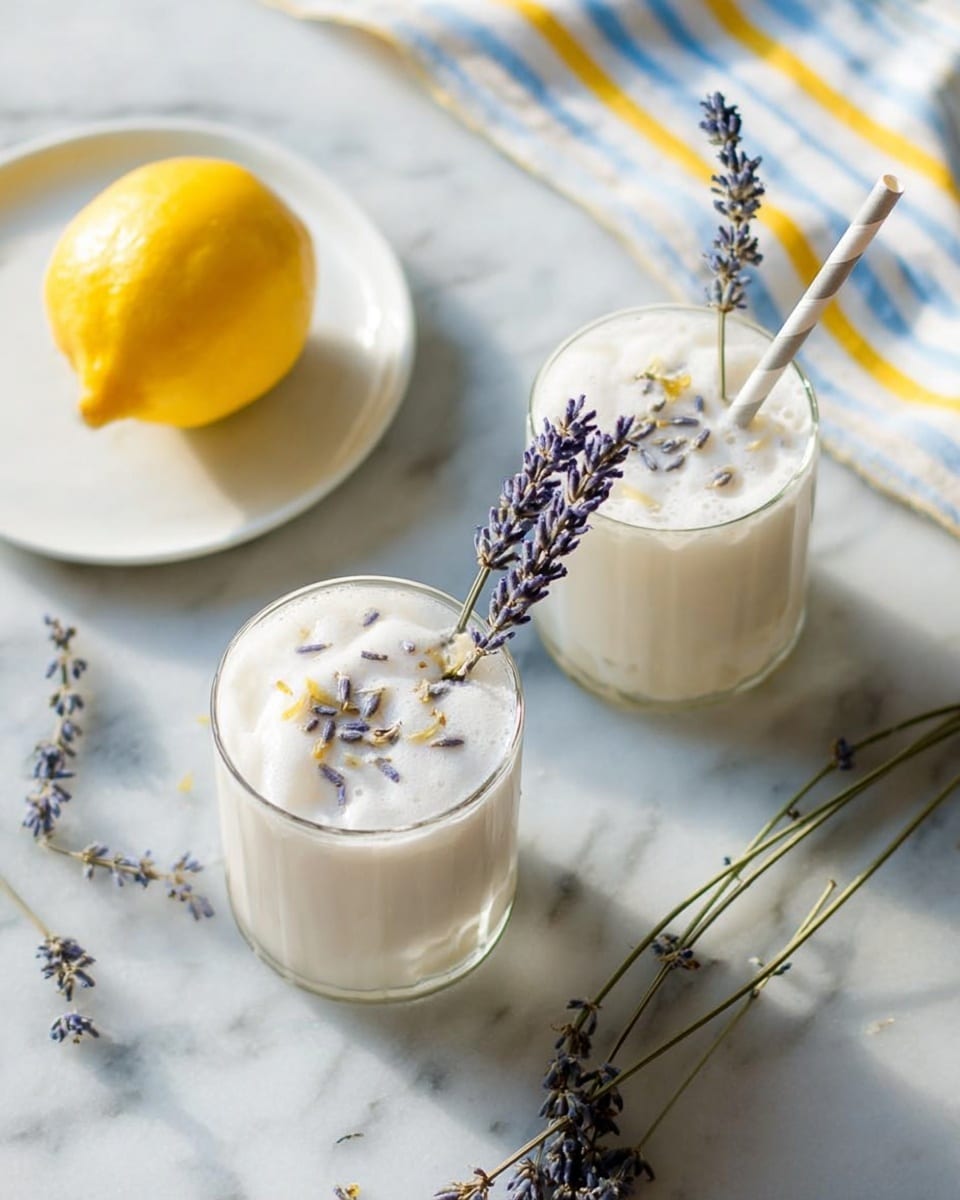Two clear glasses filled with a creamy white foam drink topped with small dried lavender buds are placed on a white marbled surface. Each glass has a striped paper straw and a sprig of fresh lavender standing upright inside. To the left, a white plate holds one bright yellow lemon. Some sprigs of lavender lie scattered on the surface near the drinks. In the top right corner, part of a striped fabric in soft blue and yellow tones is visible. The lighting is soft and natural. photo taken with an iphone --ar 4:5 --v 7
