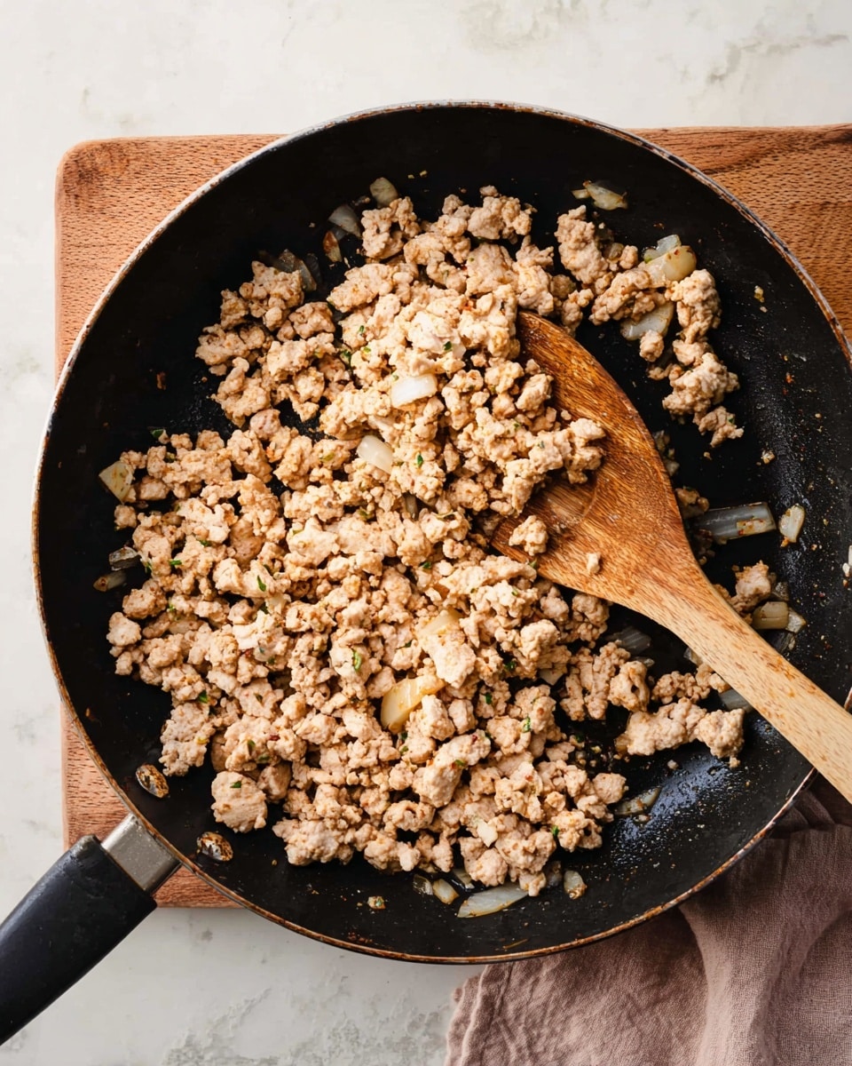 A black frying pan filled with cooked ground turkey that is light brown and beige in color, mixed with small pieces of sautéed onions. A wooden spoon with a round flat edge is resting inside the pan, partially lifting some of the turkey. The pan is set on a wooden cutting board and placed on a white marbled surface. Photo taken with an iphone --ar 4:5 --v 7