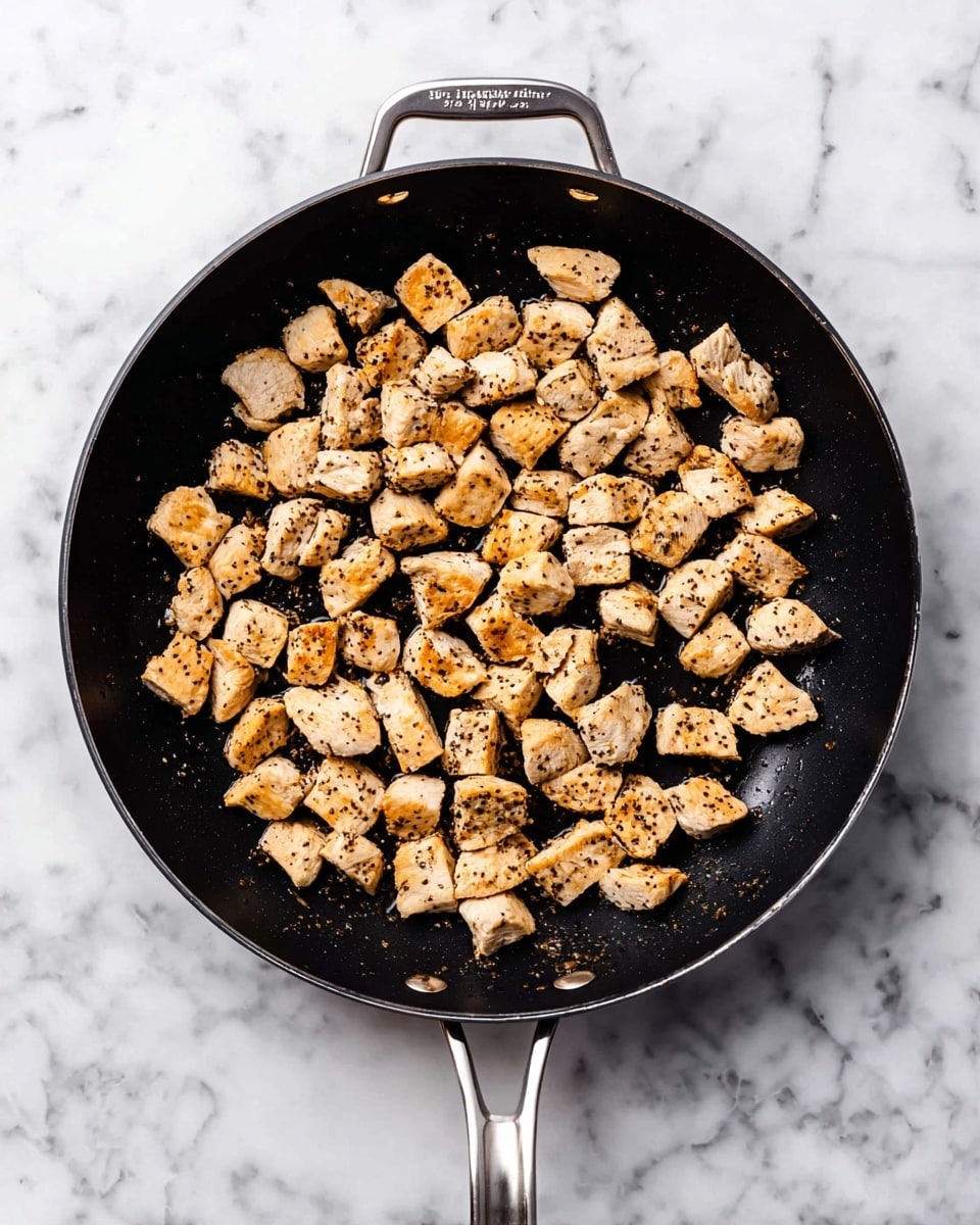 In the image, a black frying pan with silver handles sits on a white marbled surface. Inside the pan are many small pieces of cooked chicken, scattered evenly across the pan. The chicken pieces have a light golden-brown color with specks of black pepper visible on top. The texture looks tender with slightly browned edges, and the pan has a slight shine suggesting it is hot or lightly oiled. The scene is clean and bright, focusing on the pan and its cooked chicken pieces photo taken with an iphone --ar 4:5 --v 7