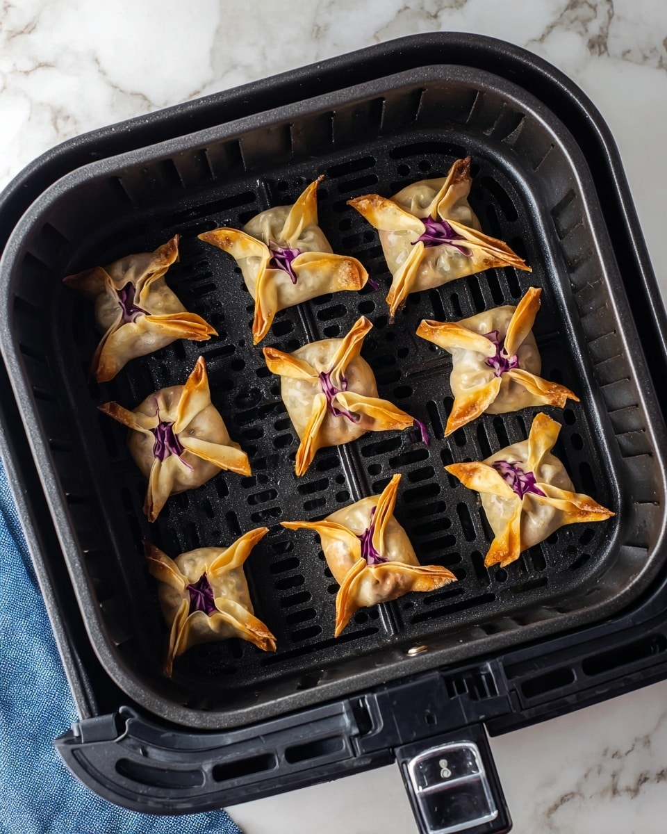 The image shows eight small square-shaped dumplings inside a black air fryer basket with a textured grid pattern. Each dumpling has a golden-brown edge and a peel in the middle that is a mix of light beige and dark purple colors. The purple parts look like cabbage leaves folded into the dough. The basket sits on a surface with a white marbled texture and a bit of a blue cloth visible at the bottom left corner. Photo taken with an iphone --ar 4:5 --v 7
