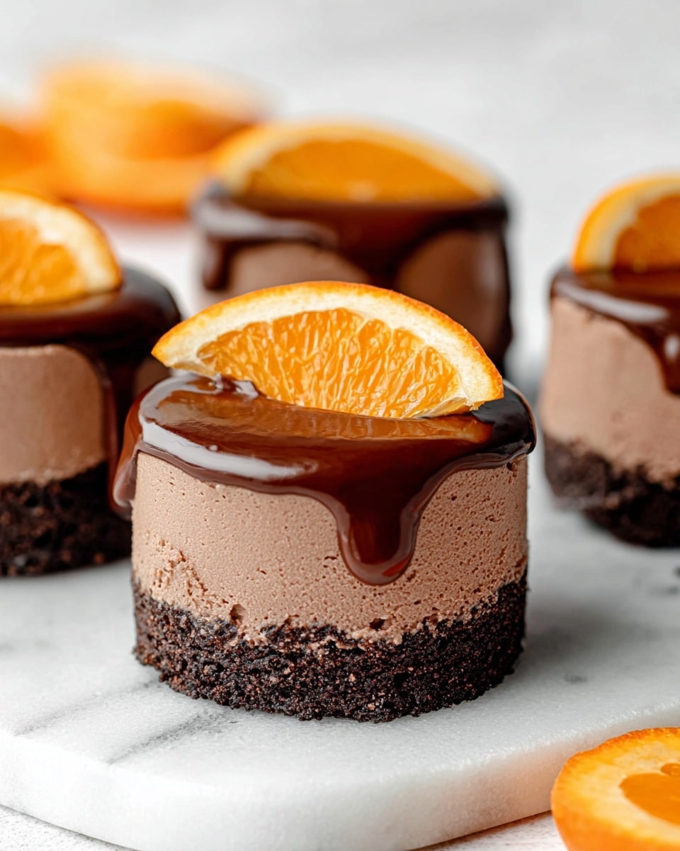 The image shows four small round chocolate cakes on a white marbled surface. Each cake has three layers: a dark brown crumbly base, a thick middle layer of smooth light brown chocolate mousse, and a glossy dark chocolate ganache dripping slightly down the sides. On top of the closest cake is a thin, curved slice of bright orange, adding a fresh pop of color. The background is soft and slightly blurred, with another orange slice visible in the distance. The cakes look rich and smooth, with the shiny ganache catching the light. photo taken with an iphone --ar 4:5 --v 7