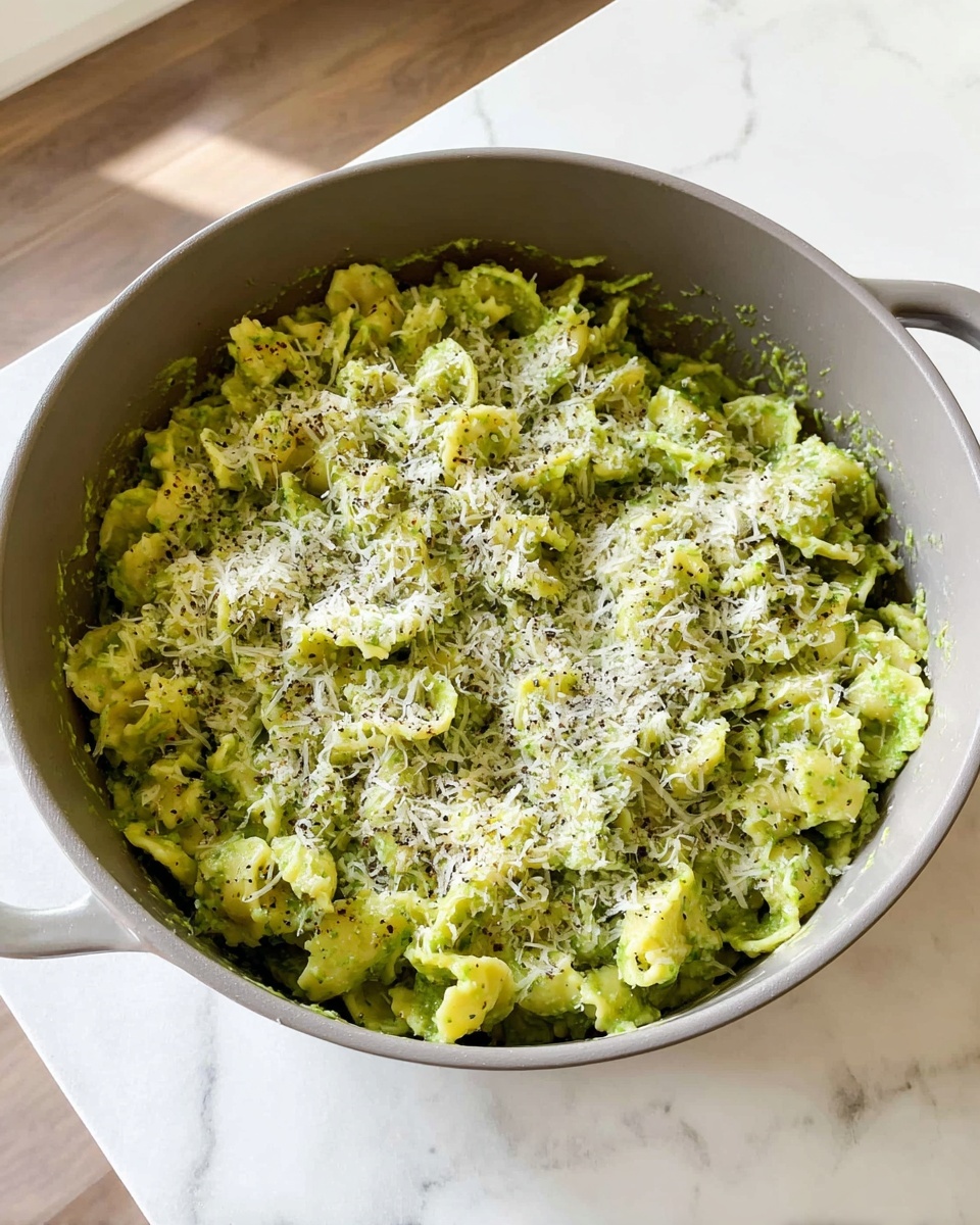 A gray pot filled with green pasta that has a curly edge. The pasta is covered evenly with grated white cheese and sprinkled with black pepper all over. The pot is sitting on a white marbled surface, with a clean, bright background showing a little wooden floor and white wall. The sauce looks creamy and thick, coating the pasta well. photo taken with an iphone --ar 4:5 --v 7