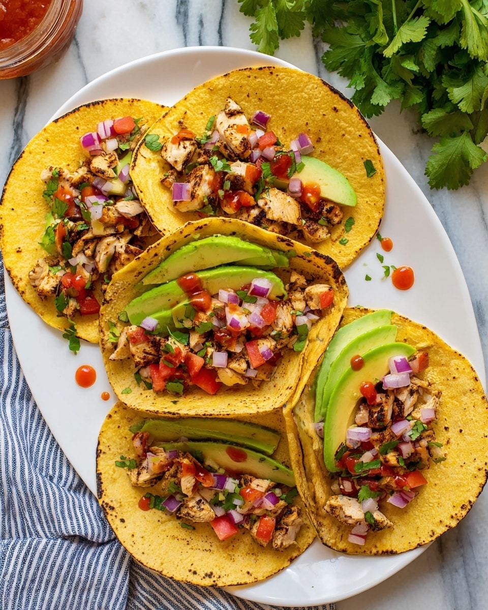 Five soft yellow corn tortillas with a light charred texture lie flat on a white plate. Each tortilla is layered with bright green sliced avocado arranged in small overlapping segments near the center, topped with small chunks of grilled light brown and slightly charred chicken. Scattered finely diced red onions and small red tomato pieces add pops of color, and small green cilantro leaves are sprinkled on top along with a few drops of red hot sauce. The plate rests on a white marbled surface with a striped blue and white cloth nearby, and fresh cilantro leaves are visible on the side. Photo taken with an iphone --ar 4:5 --v 7
