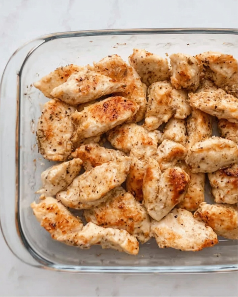 This image shows a clear glass baking dish filled with about fifteen small pieces of cooked seasoned chicken. The chicken pieces are light golden brown with some darker spots where they are cooked more, and you can see some small black pepper flakes on the surface. The chicken pieces are arranged close together, covering the bottom of the dish, and the edges of the glass dish are visible around the chicken. The background is a white marbled surface. photo taken with an iphone --ar 4:5 --v 7
