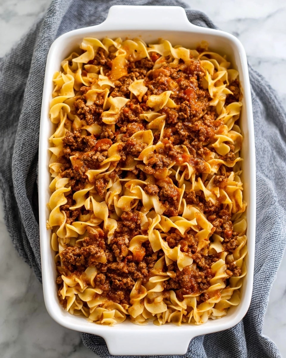 The image shows a white rectangular baking dish filled with a pasta bake dish. The dish contains two visible layers: the bottom layer has egg noodles that are pale yellow with a slight shine, while the top layer features ground meat cooked with tomato sauce, giving it a rich brown and red mixed color. The meat and noodles are mixed evenly, creating a textured surface with twisted noodles and small meat chunks scattered throughout. The baking dish is placed on a soft gray cloth over a white marbled surface. photo taken with an iphone --ar 4:5 --v 7