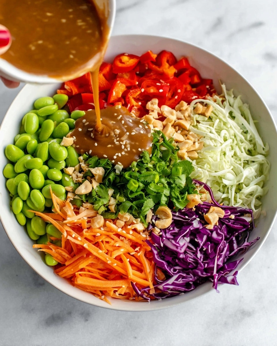 A white bowl on a white marbled surface holds chopped red bell peppers with sesame and peanuts on the bottom left, bright green edamame beans with sesame and peanuts on the bottom right, shredded orange carrots with peanuts above the edamame, chopped green onions at the top right, chopped fresh green cilantro in the center, shredded white cabbage with peanuts at the top left, and shredded purple cabbage with peanuts just below the white cabbage. A woman's hand with red nail polish pours thick brown dressing over the center cilantro. photo taken with an iphone --ar 4:5 --v 7