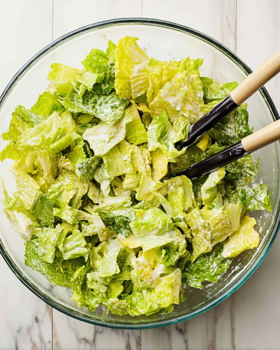 A clear glass bowl filled with chopped, mixed green lettuce leaves, ranging from bright yellow-green to darker green shades, all lightly coated in creamy white dressing. Two black and beige salad tongs rest inside the bowl, with their ends dipped in the lettuce mix, sitting on a white marbled surface. The lettuce pieces are uneven but fresh-looking, giving a textured, leafy appearance. Photo taken with an iphone --ar 4:5 --v 7