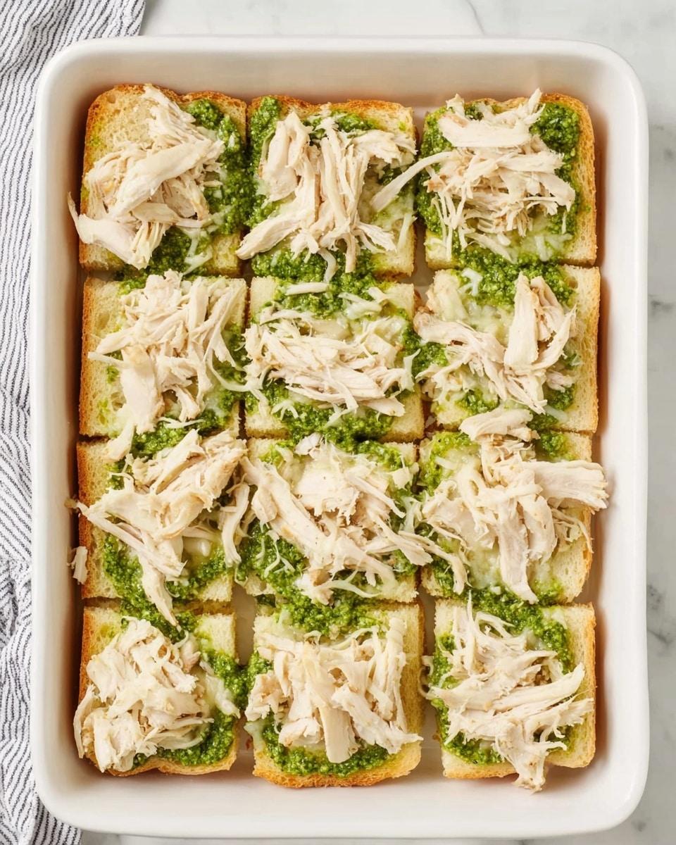 The image shows a white rectangular dish filled with a layer of twelve square bread pieces arranged in a 3 by 4 grid. Each piece of bread has a spread of bright green pesto layered on top, with shredded light beige chicken pieces scattered over the pesto, creating a textured and layered look. The background surface is white marble, and a striped towel is partially visible on the left side of the dish. Photo taken with an iphone --ar 4:5 --v 7