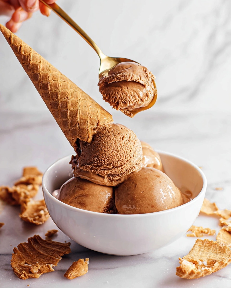 A white bowl is filled with three scoops of rich brown ice cream, with a light shine on its creamy texture. A waffle cone is placed upside down on top of the ice cream, slightly melting the scoop beneath it. A woman's hand holds a gold spoon with a small scoop of the same ice cream above the bowl. The bowl sits on a white marbled surface, surrounded by broken waffle cone pieces scattered around. Photo taken with an iphone --ar 4:5 --v 7