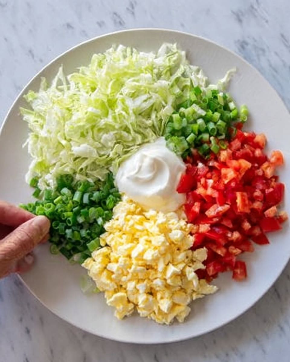 The image shows a white plate on a white marbled surface with four colorful layers of chopped ingredients arranged in sections around the plate. At the top, there is a pile of shredded green lettuce. To the right of the lettuce, there are small, diced red tomatoes. Below the tomatoes, finely chopped green onions or scallions are placed. To the left and near the center bottom, there is a mound of finely chopped yellow hard-boiled eggs. At the center of the plate, there is a dollop of white creamy mayonnaise. A woman's hand is reaching towards the plate from the top left corner. Photo taken with an iphone --ar 4:5 --v 7