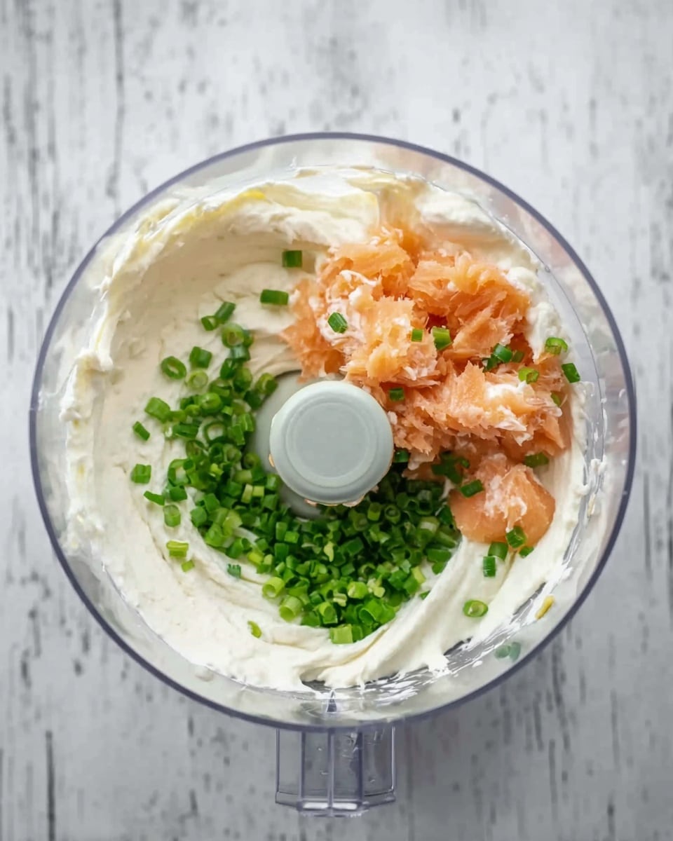 A clear food processor bowl holds three visible layers: a thick white creamy base spread evenly inside the bowl, small pieces of light orange fish placed on one side, and finely chopped green onions positioned opposite the fish near the center blade. The bowl sits on a white marbled surface. Photo taken with an iphone --ar 4:5 --v 7