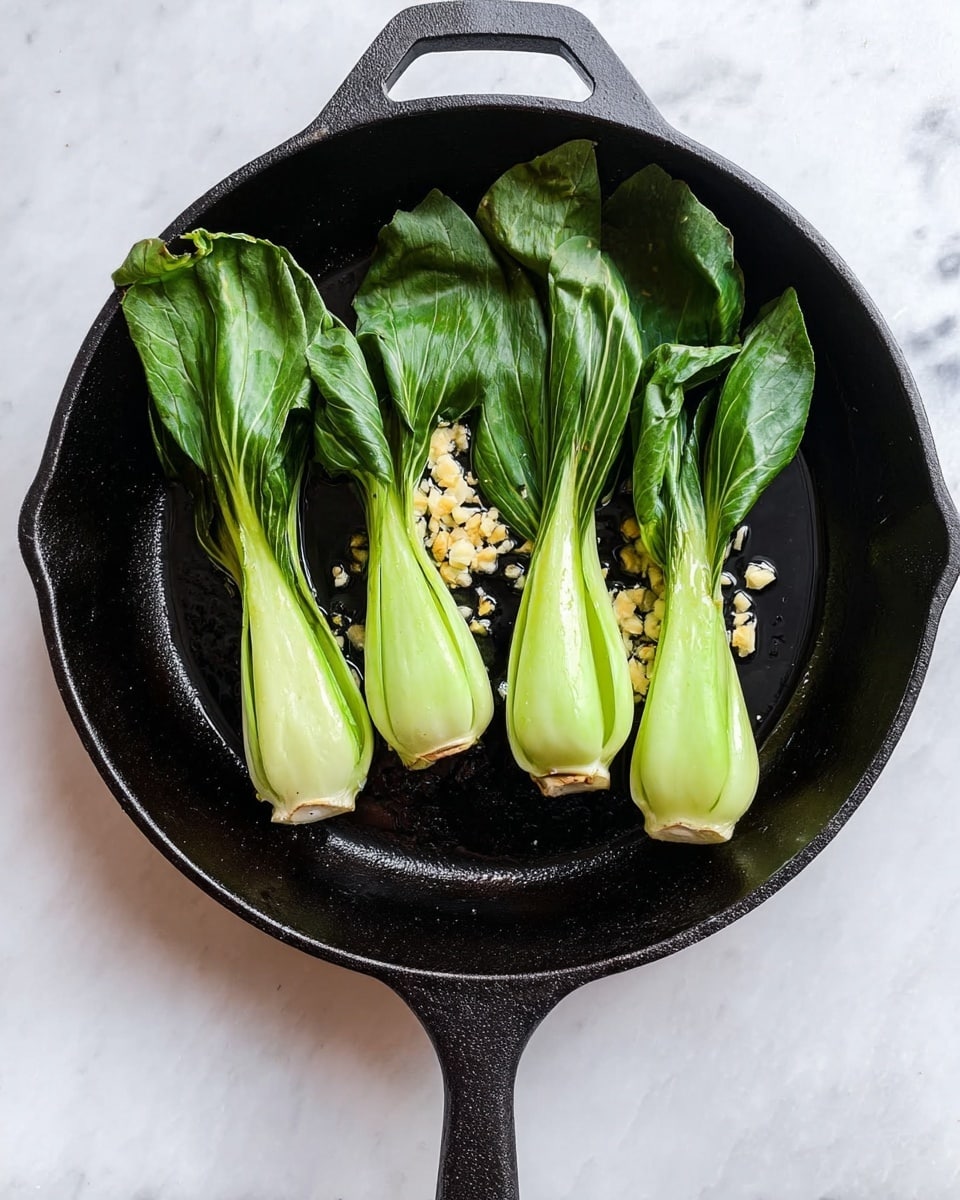 A black cast iron skillet sits on a white marbled surface, holding four whole bok choy arranged side by side with their light green bulbs at the bottom and darker green leaves pointing upward. Small pieces of chopped garlic scatter in the center beneath the bok choy, contrasting with the smooth skillet surface. The skillet handle extends downwards from the bottom center of the image. photo taken with an iphone --ar 4:5 --v 7