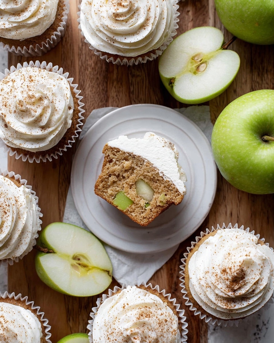 The image shows a close-up of several cupcakes with white frosting lightly dusted with brown cinnamon powder on top. One cupcake is open on a white plate, showing the inside with visible small pieces of green apple embedded in the light brown cake. Around the cupcakes, there are whole green apples and halved green apples with a bright green skin and white inside, all placed on a wooden surface. The cupcakes have white paper liners, and the frosting is fluffy and swirled. The composition is neat, with the cupcakes and apples evenly spread around. The background is changed to a white marbled texture. photo taken with an iphone --ar 4:5 --v 7