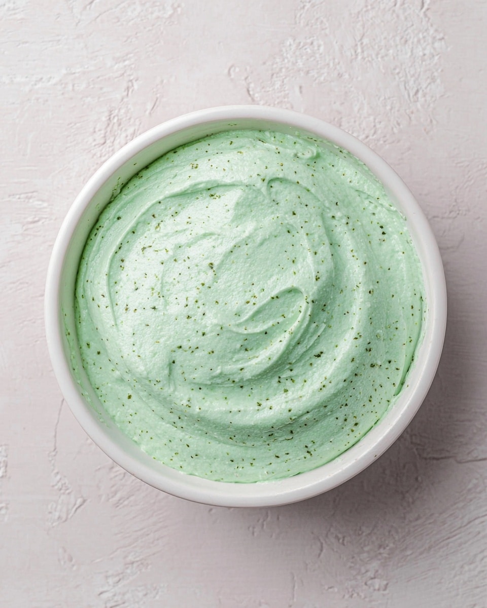 A white bowl filled with one smooth layer of creamy dip in pale green color with tiny darker green flecks spread evenly across its surface positioned on a white marbled texture background photo taken with an iphone --ar 4:5 --v 7