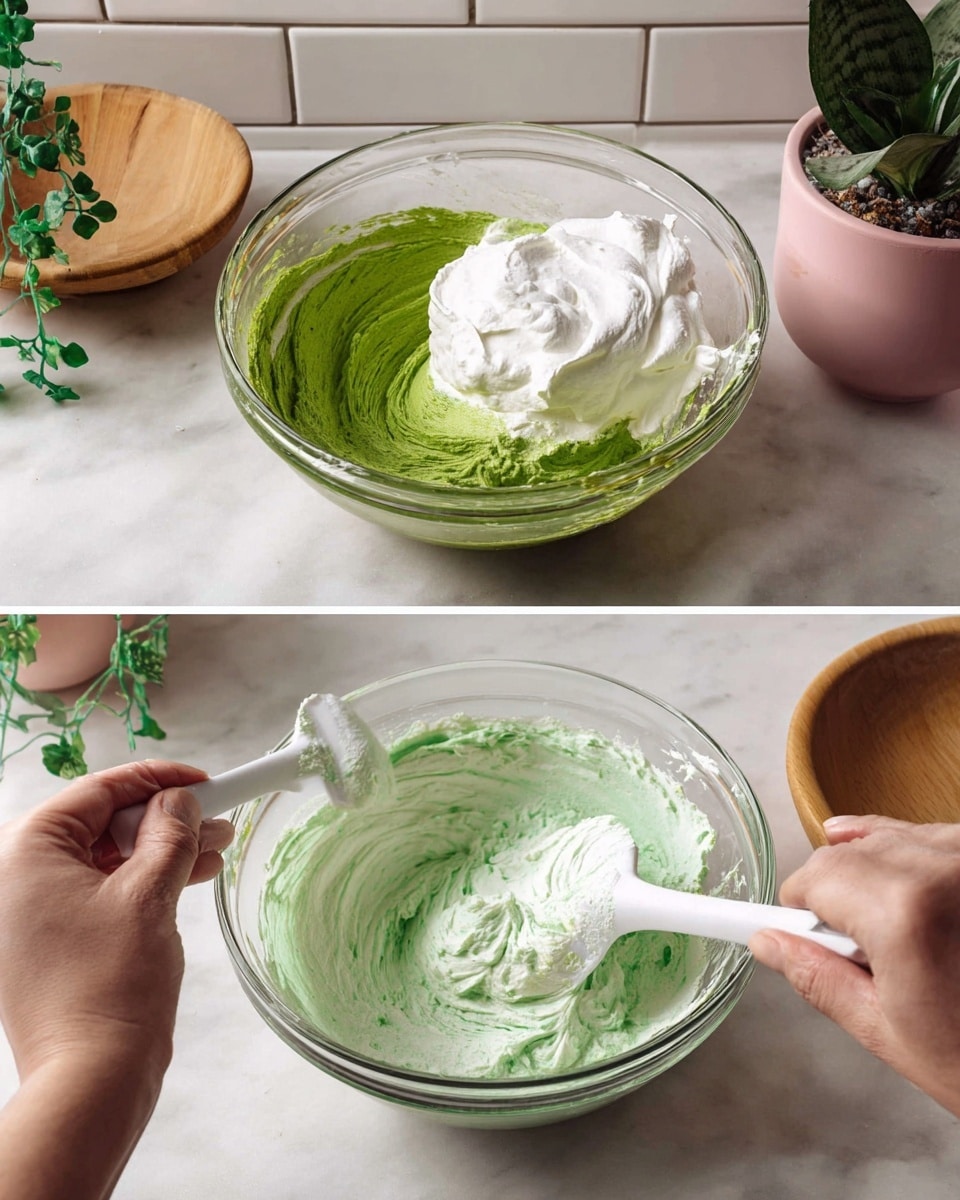 The image shows two clear glass bowls on a white marbled surface with white tiled wall in the background. The bowl on the left contains a thick green mixture, with a large white fluffy layer of whipped cream being folded into it using a white spatula held by a woman's hand. The bowl on the right has a mix of the green and white ingredients stirred together, creating a light green creamy texture with splotches of deeper green. A woman’s hand stirs the mixture with the same white spatula. A pink pot with green hanging plants and a wooden bowl are visible in the background. Photo taken with an iphone --ar 4:5 --v 7