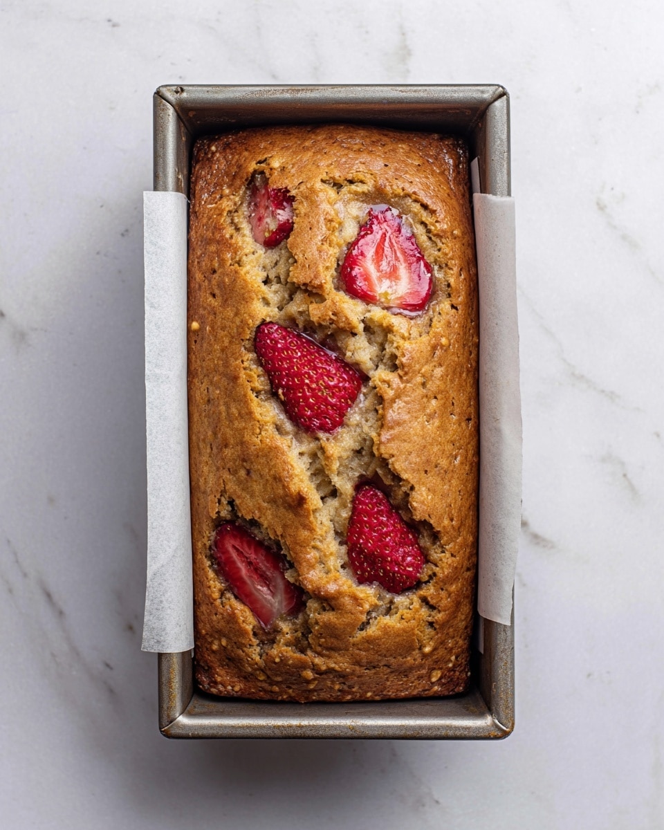 The image shows a freshly baked loaf of bread in a rectangular metal pan lined with white parchment paper. The top of the loaf is golden brown with a slightly rough texture and has four large slices of red strawberries embedded on the surface, providing a pop of color. The pan sits on a white marbled surface, which adds a clean and bright contrast to the warm tones of the bread. The bread looks moist and dense with some cracks on the top showing its soft inside. photo taken with an iphone --ar 4:5 --v 7