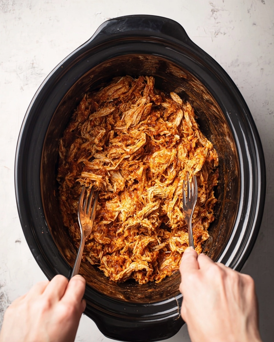 Inside a round black slow cooker, there is one layer of shredded chicken mixed with reddish-orange sauce, giving it a textured and moist look. Two silver forks, each held by a woman’s hand from opposite sides, are pulling the chicken apart as it sits against the smooth black inner surface of the cooker. The cooker is placed on a white marbled surface that can be partially seen around the edges. The overall image shows a close-up view focused on the cooker’s interior and the shredded chicken being pulled apart. photo taken with an iphone --ar 4:5 --v 7