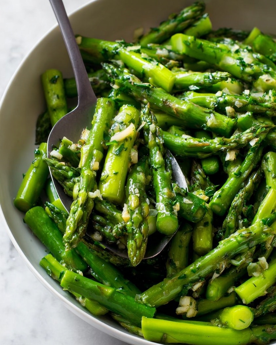 The image shows a close-up of many bright green asparagus pieces in a white bowl, all cut into short sections with some tips visible. The asparagus is mixed with bits of garlic and herbs, which look small, off-white, and green, giving the dish texture and color variety. A gray spoon is seen scooping some asparagus on the left side, its smooth surface contrasting with the rough texture of the vegetables. The shiny asparagus looks cooked and moist, catching the light to show a fresh, glossy finish. The background is a white marbled surface. photo taken with an iphone --ar 4:5 --v 7