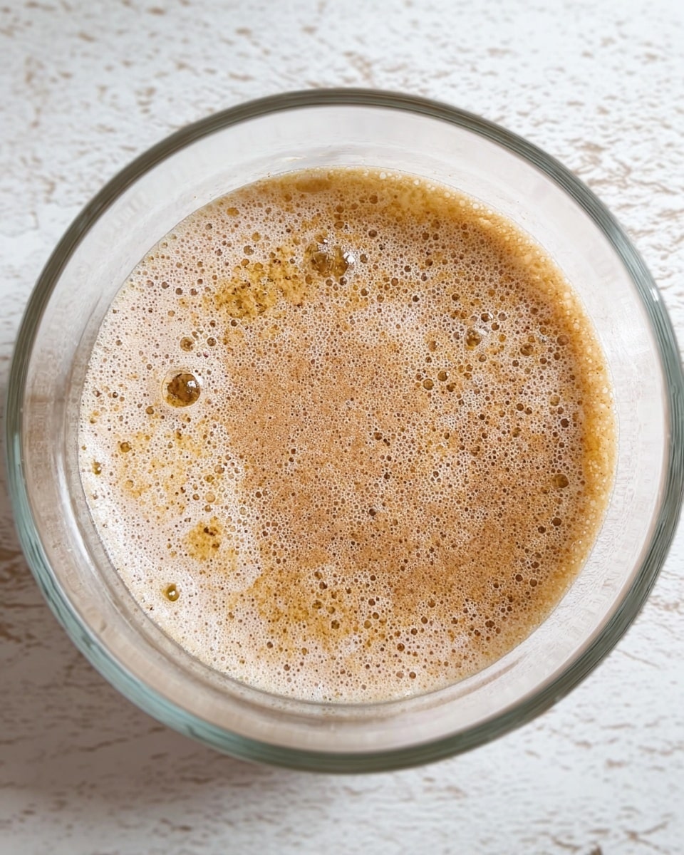 A clear glass bowl filled with a light brown frothy mixture that has small bubbles and specks floating on the surface, sitting on a white marbled textured surface. The mixture appears to have a creamy texture with a layer of light brown powdery specks evenly spread throughout. The bowl is round and transparent, showing the mixture clearly inside. Photo taken with an iphone --ar 4:5 --v 7