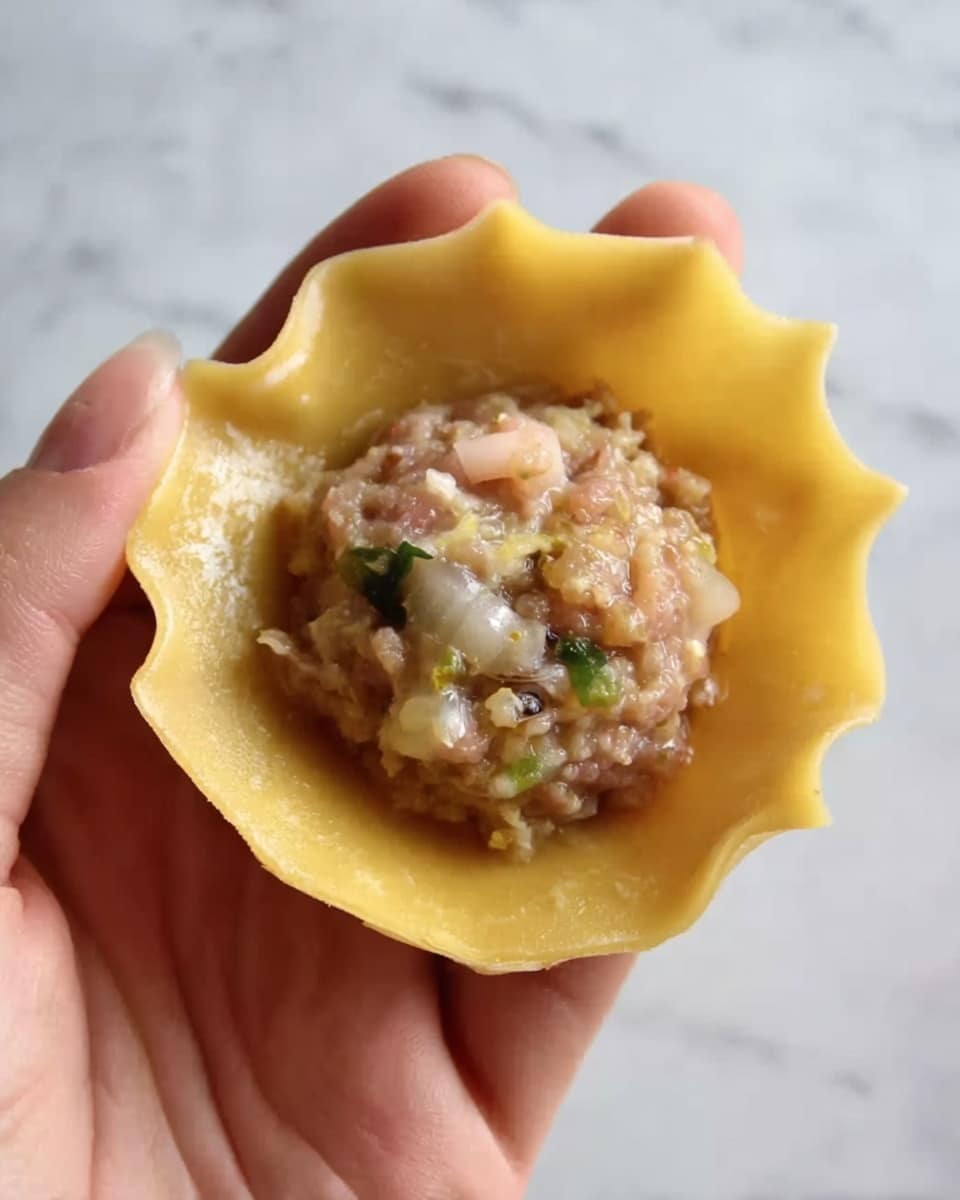 A woman’s hand holds a single thin, round, yellow dough wrapper with slightly uneven edges. In the center of the dough is a small mound of raw mixed minced filling that has a pinkish-gray color with small bits of green, white, and translucent ingredients visible. The background features a white marbled texture. photo taken with an iphone --ar 4:5 --v 7