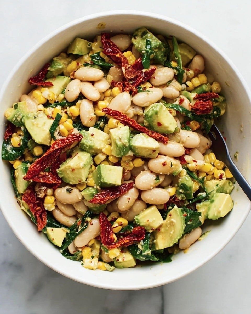 A white bowl filled with a colorful salad, showing several layers and textures mixed together. The salad contains beige white beans that are oval and smooth, yellow corn pieces cut in small rounds with a slight char, bright green diced avocado cubes, dark green leafy vegetable pieces, and strips of red sun-dried tomatoes scattered around. The salad looks fresh with a mix of soft and slightly firm textures. A black spoon is partially visible inside the bowl. The bowl is placed on a white marbled surface. Photo taken with an iphone --ar 4:5 --v 7