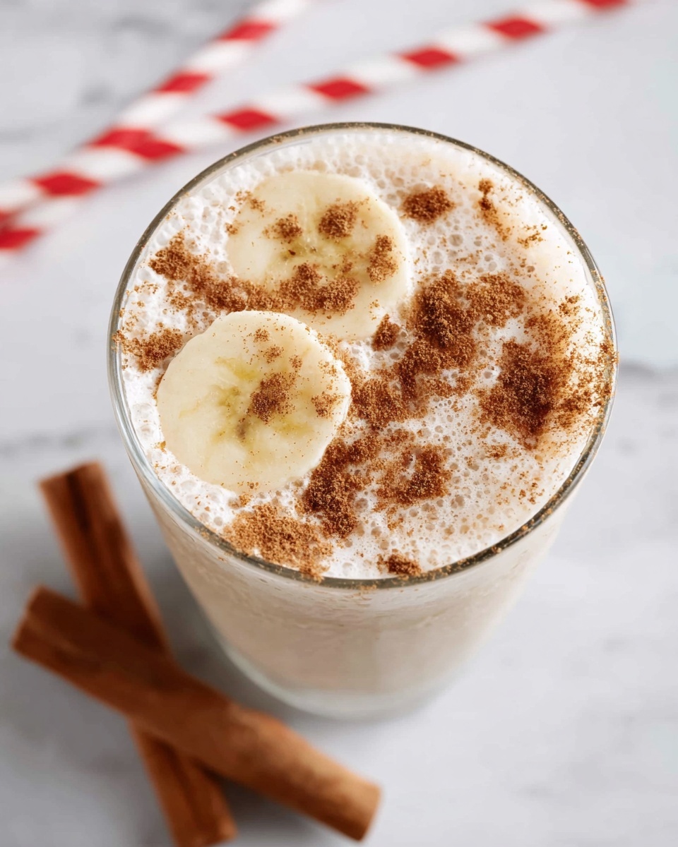 A close-up view of a glass filled with a creamy, light beige drink, topped with two banana slices partially submerged in the frothy surface. The creamy top is sprinkled unevenly with brown cinnamon powder, adding a speckled texture. The glass sits on a white marbled surface with a blurred red and white striped straw laid diagonally in the background and two cinnamon sticks placed nearby. The overall look is fresh and inviting, with soft lighting that highlights the creaminess and texture of the drink. photo taken with an iphone --ar 4:5 --v 7