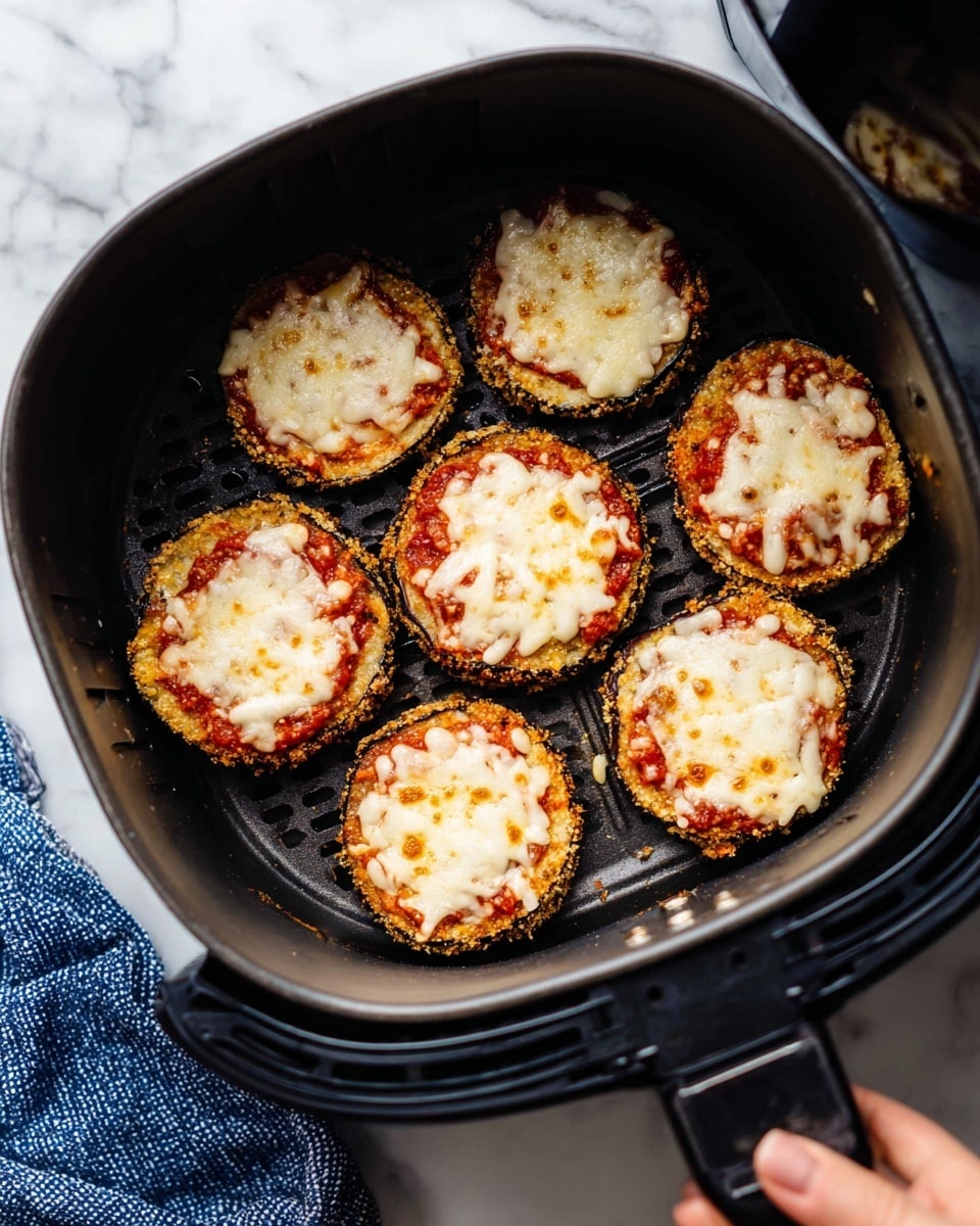 The image shows seven small round eggplant slices arranged in a single layer inside a dark air fryer basket. Each eggplant slice has a crispy golden-brown breaded edge, a middle layer of red tomato sauce, and a top layer of melted white cheese with light golden-brown spots. The air fryer basket is being held by a woman's hand and sits on a white marbled surface with a blue textured cloth beneath it. photo taken with an iphone --ar 4:5 --v 7