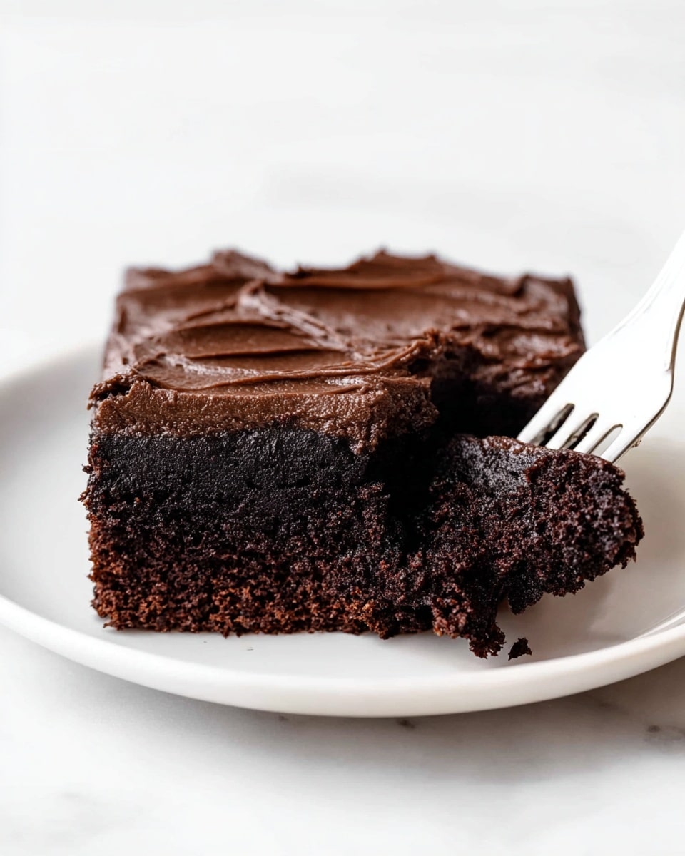 A square piece of dark brown chocolate cake sits in the center of a white plate on a white marbled surface. The cake has two layers: a dense, moist bottom layer with a slightly crumbly texture and a thick, smooth layer of dark chocolate frosting on top, featuring swirled patterns. A white plastic fork pokes into the front right corner of the cake, lifting a small chunk, showing the rich, soft inside. The overall scene is bright and clean, focused closely on the cake and fork. photo taken with an iphone --ar 4:5 --v 7