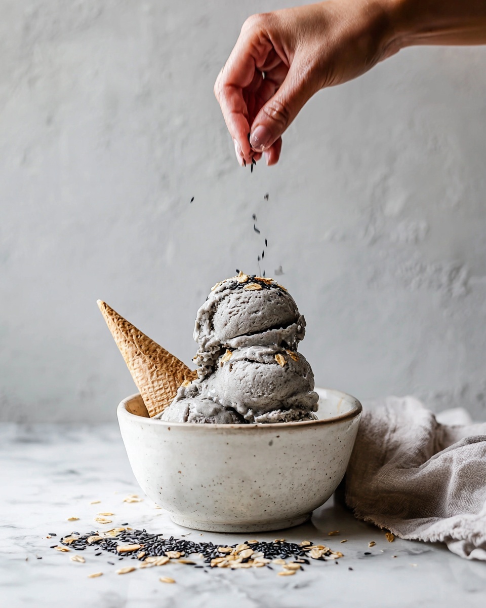 A white ceramic bowl filled with three scoops of grayish ice cream stacked on top of each other. The ice cream has a rough texture with some golden flakes scattered on it. A light beige cone is placed leaning inside the bowl on the left side. Crushed flakes and some black seeds are spread in front of the bowl on a white marbled surface. A woman's hand is seen above the ice cream, sprinkling small black seeds over the top scoop. A soft, light-colored cloth is draped to the right side of the bowl. The background is a simple light gray textured wall. Photo taken with an iphone --ar 4:5 --v 7
