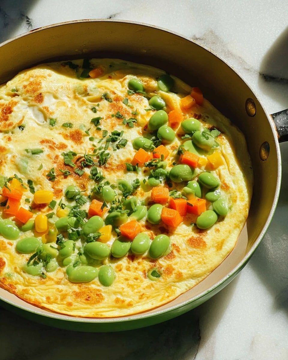 A cooked omelette sits in a green pan, its surface golden yellow with light brown spots. The omelette has three main layers: a smooth, pale yellow egg base, scattered bright green edamame beans, and small cubes of orange carrots mixed with some yellow corn kernels and chopped green herbs on top. The pan is on a white marbled surface with sunlight casting soft shadows. Photo taken with an iphone --ar 4:5 --v 7