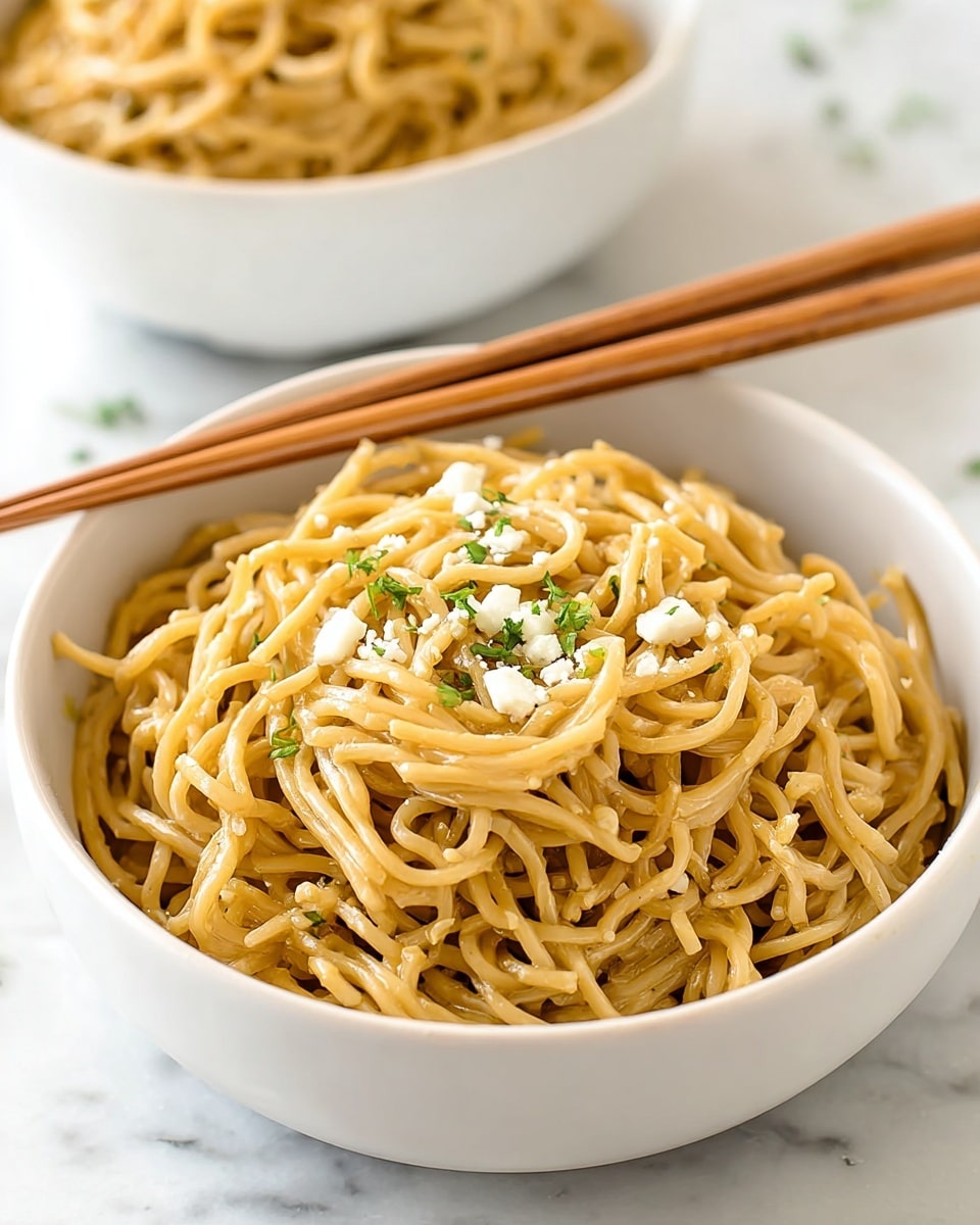 A white bowl filled with a mound of light brown noodles, topped with small pieces of white cheese and tiny green herb bits scattered evenly on top and mixed lightly through the noodles. The noodles appear soft and slightly shiny. Resting on the edge of the bowl is a pair of light brown wooden chopsticks. The bowl is placed on a white marbled surface, with another similar bowl slightly out of focus in the background. photo taken with an iphone --ar 4:5 --v 7
