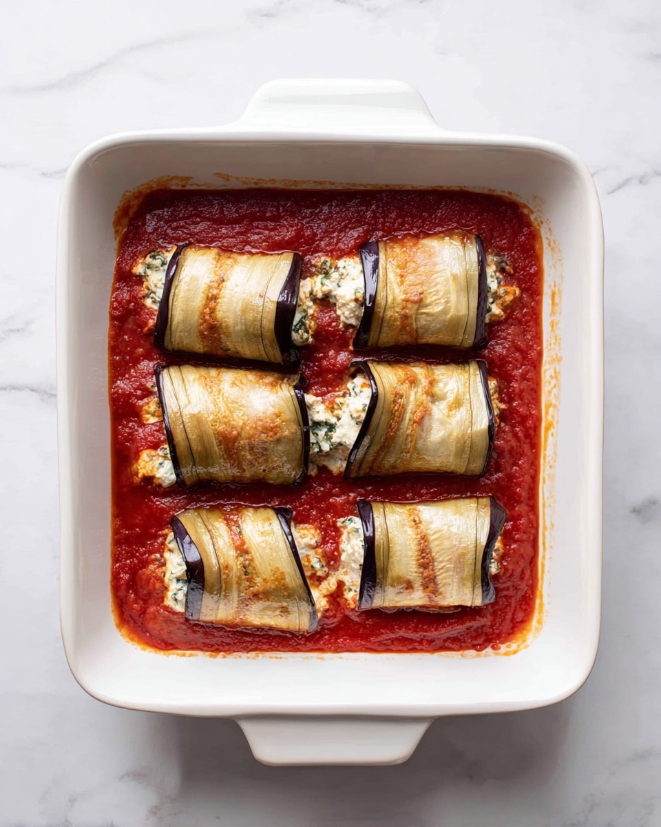 The image shows a white square baking dish filled with a layer of thick red tomato sauce covering the bottom. On top, there are six rolled pieces of lightly cooked eggplant arranged in two rows of three. Each rolled eggplant slice is light brown and purple with a soft, slightly wrinkled texture, filled with a creamy white mixture that peeks out from the sides, likely cheese or a ricotta mix with some green herbs. The dish sits on a white marbled surface. Photo taken with an iphone --ar 4:5 --v 7