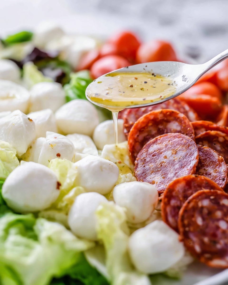The image shows a close-up of a white plate with a layered snack arrangement on a white marbled surface. The base layer is fresh green lettuce leaves, topped with small white mozzarella balls scattered across. To the right, there is a neat pile of red pepperoni slices with visible pepper flakes. Above the mozzarella, a spoon holds a golden yellow dressing with black pepper specks dripping down onto the mozzarella. The background is softly blurred, highlighting the fresh and colorful ingredients. Photo taken with an iphone --ar 4:5 --v 7