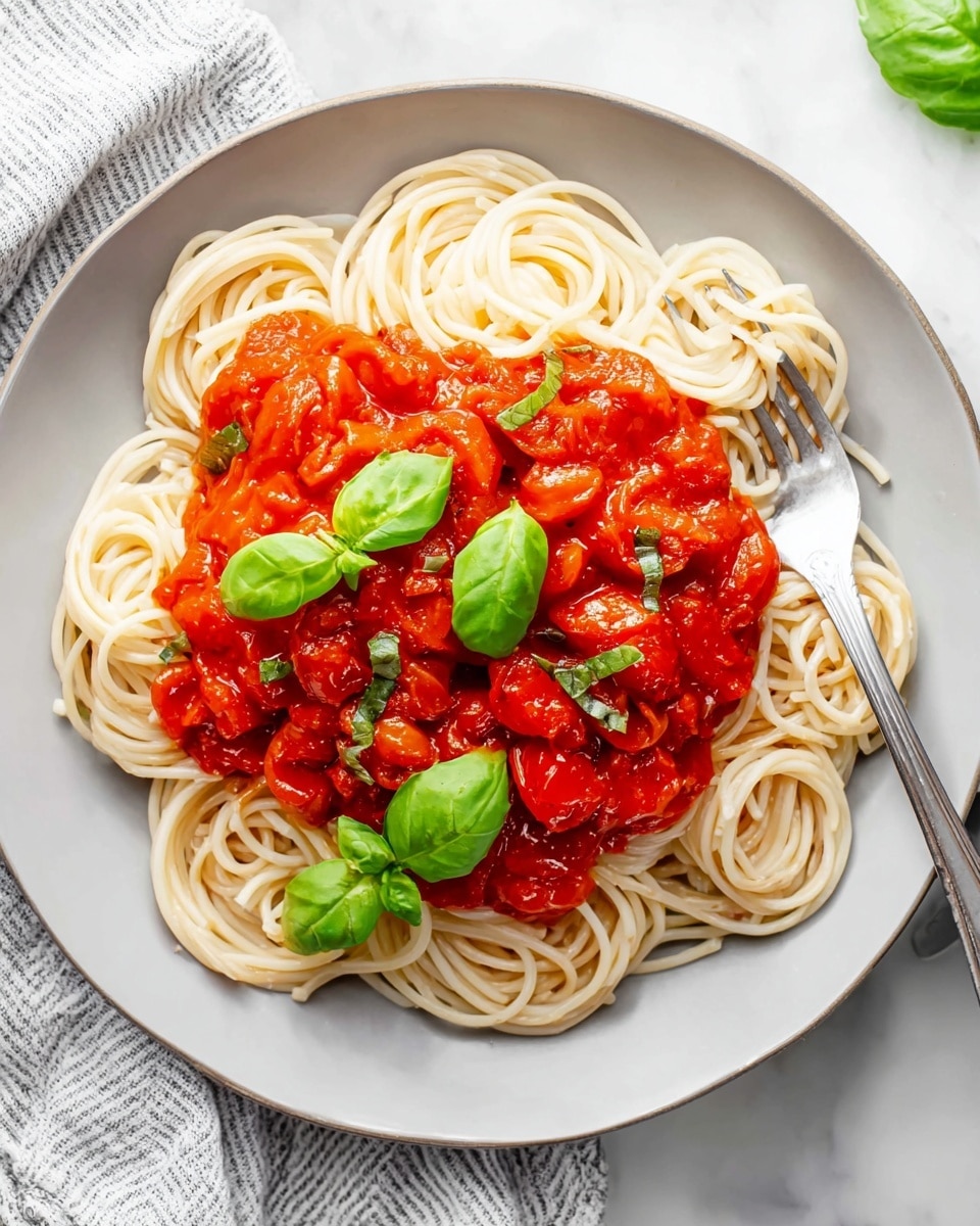 A white plate holds a neat round layer of light beige spaghetti noodles as the base. On top lies a thick, bright red tomato sauce with visible chunks of cooked tomatoes mixed with small green herb pieces scattered throughout. Fresh green basil leaves are placed evenly across the sauce, adding a pop of color. A silver fork rests on the right side of the plate, partially inserted into the noodles. The plate sits on a white marbled surface, with a soft striped cloth nearby. Photo taken with an iphone --ar 4:5 --v 7