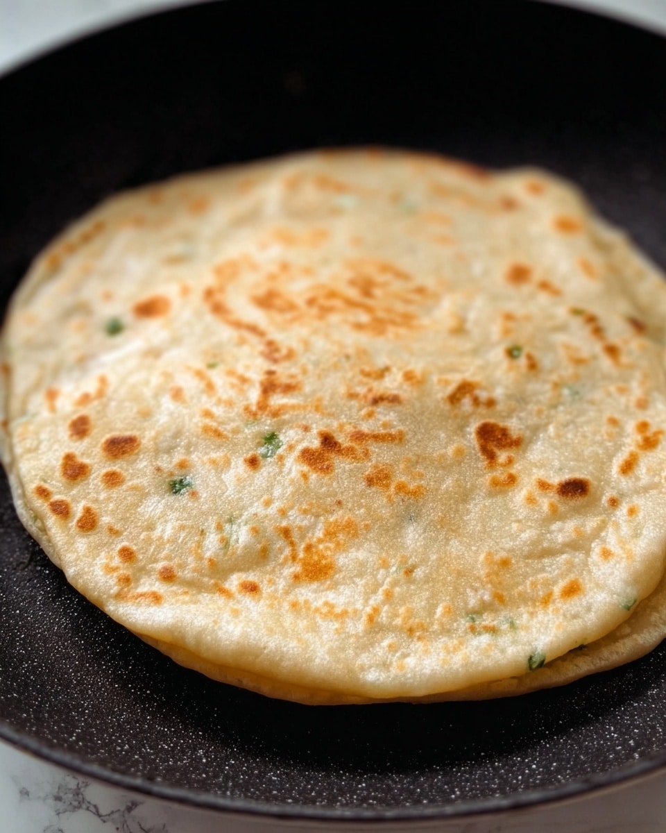 The image shows a single round flatbread puffed up in the middle while cooking on a black pan. The flatbread is light beige with small and scattered golden-brown spots all over its surface, and tiny green specks are visible inside the dough. The flatbread looks soft and slightly fluffy, with thin edges that are pressed and sealed, giving it a stuffed appearance. The pan has a smooth dark surface, and the photo captures a close-up side view of the bread. The background is a white marbled texture. Photo taken with an iphone --ar 4:5 --v 7
