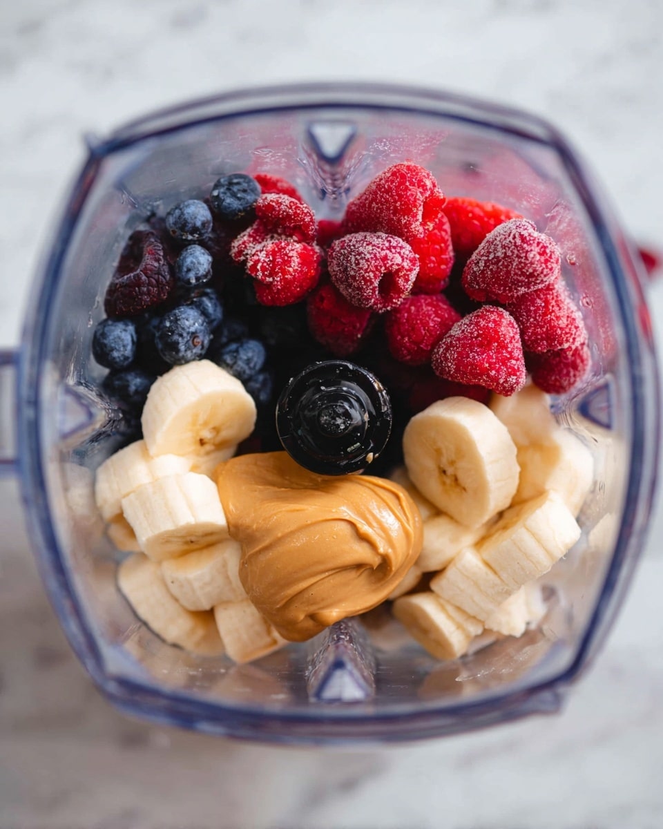 Inside a clear blender, the bottom layer shows a mix of blueberries and strawberries with a deep blue and bright red color. Next, on top, there is a thick dollop of creamy peanut butter in golden brown. Around the peanut butter, several banana slices spread out in light yellow, mostly frozen with a frosty texture. Scattered frozen raspberries add pops of red on the top, giving a fresh look. The blender blades and central black mechanism are visible in the center amidst the fruits. The whole scene is shot on a white marbled surface. photo taken with an iphone --ar 4:5 --v 7