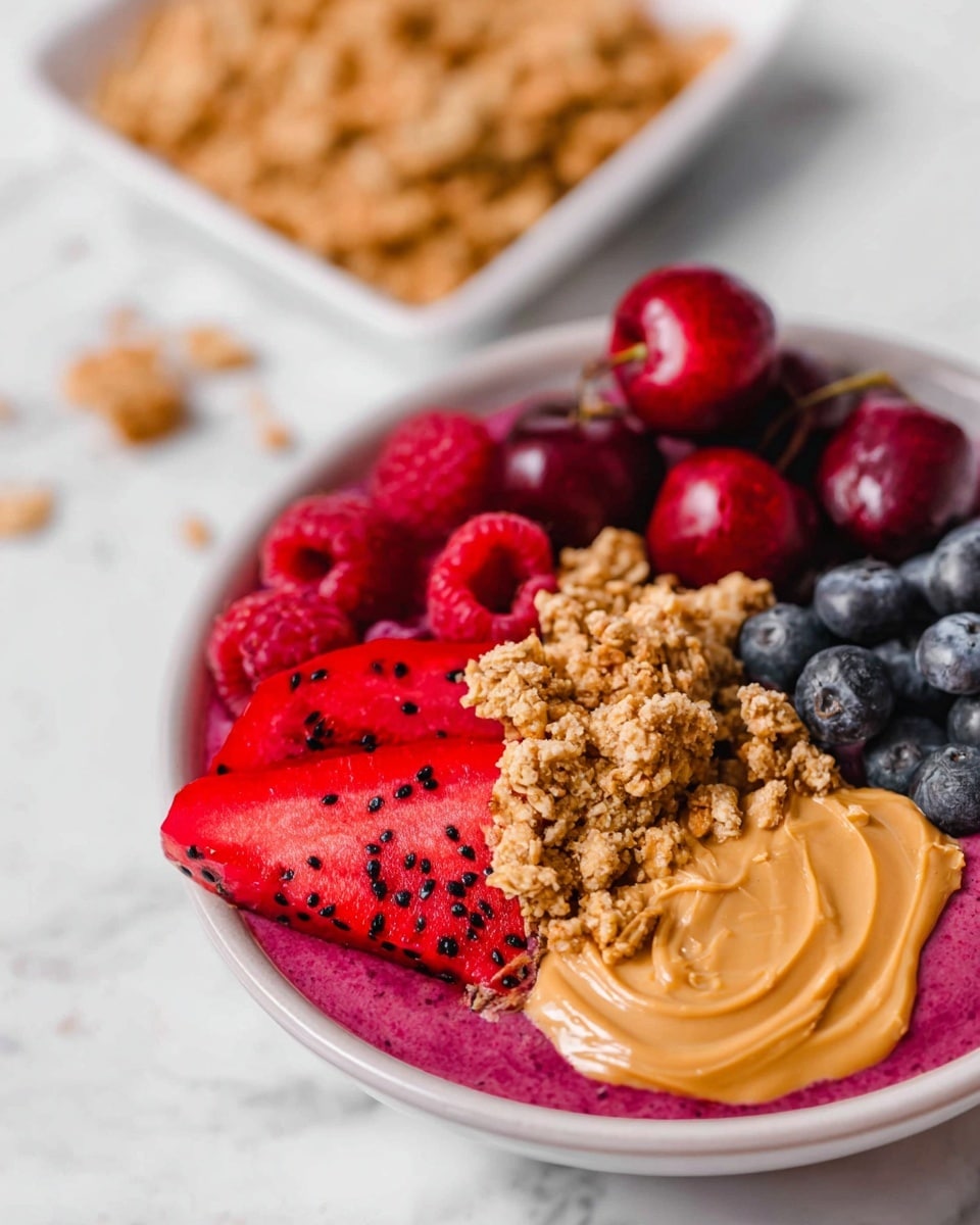 A white bowl shows a colorful mix of food. The bottom layer is a deep pink smoothie with a thick texture. On top, there are five main layers arranged in sections: bright red raspberries on the left, dark red halved cherries next to them, purple blueberries towards the back, a pile of light brown crunchy granola in the center, and a smooth swirl of light brown peanut butter on the right side. In front of the peanut butter, there are a few slices of bright red dragon fruit with black seeds. The bowl sits on a white marbled surface with a white square bowl of granola blurred in the background photo taken with an iphone --ar 4:5 --v 7
