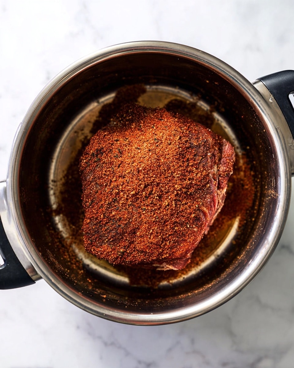 The image shows a round piece of meat inside a silver metal pot, covered with a thick layer of reddish-brown dry spice rub that has a coarse, textured look. The meat sits centered in the pot, surrounded by a small amount of clear liquid, with the metal of the pot shining smoothly around it. The pot handles are black, and the background is a white marbled texture. photo taken with an iphone --ar 4:5 --v 7