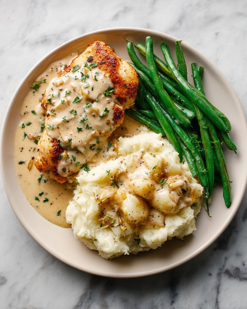 A round white plate holds a meal with three main sections neatly arranged. On the top left is a piece of golden-brown cooked chicken covered with a creamy, light beige sauce sprinkled with small green herbs. To the right of the chicken, there is a large scoop of creamy mashed potatoes with visible roasted garlic cloves and black pepper, also topped with sauce and green herbs. On the bottom left of the plate, bright green cooked green beans lie side by side, lightly drizzled with the same creamy sauce. The plate rests on a white marbled surface. photo taken with an iphone --ar 4:5 --v 7