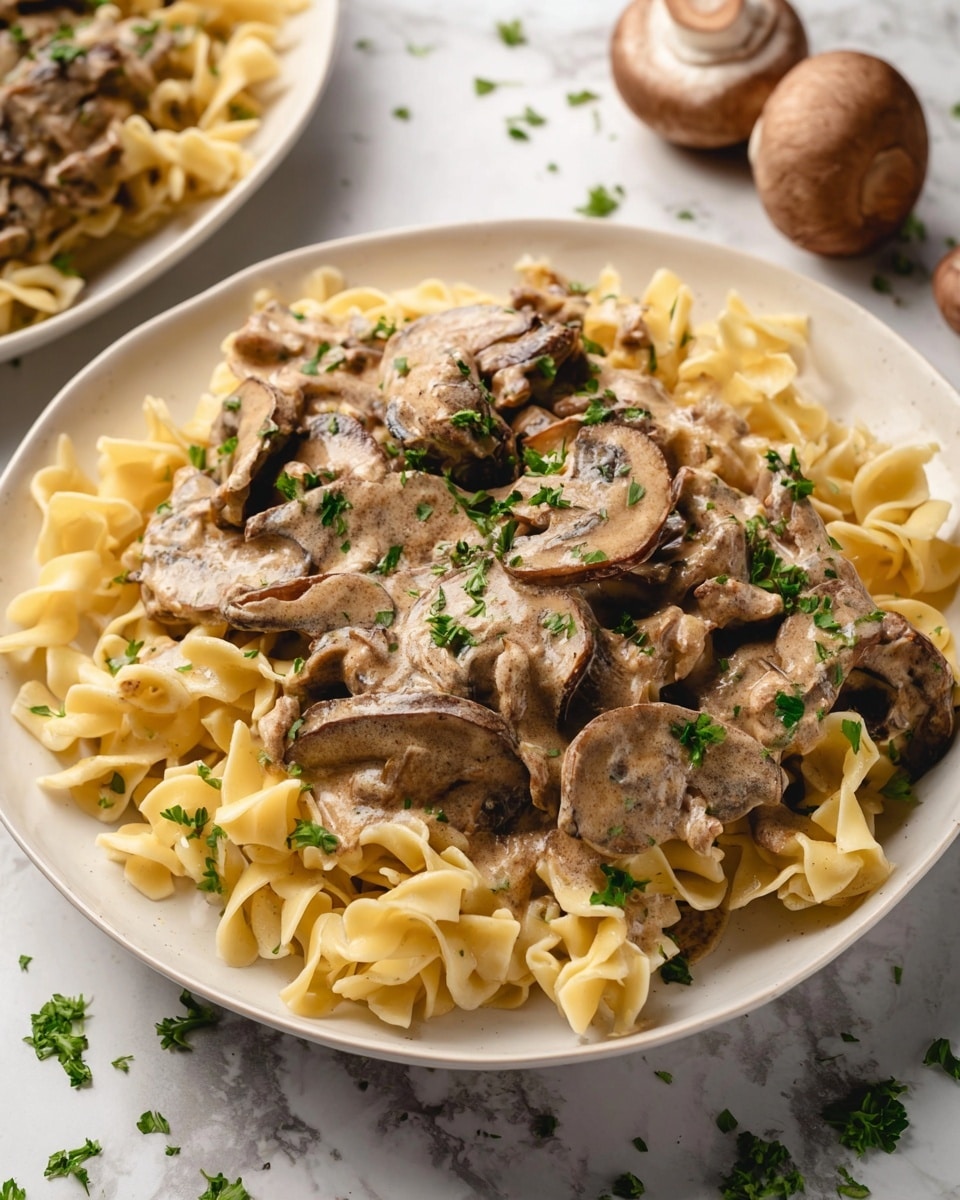 The dish shows a white plate filled with three layers: the bottom layer is pale yellow egg noodles with a soft and curly texture spread evenly; the second layer is thick slices of brown mushrooms placed on top but lightly mixed into the noodles; the top layer is a creamy, light brown sauce coating the mushrooms and noodles, garnished with small pieces of chopped green parsley sprinkled over the whole dish. The plate sits on a white marbled surface with a few whole brown mushrooms in the background. photo taken with an iphone --ar 4:5 --v 7