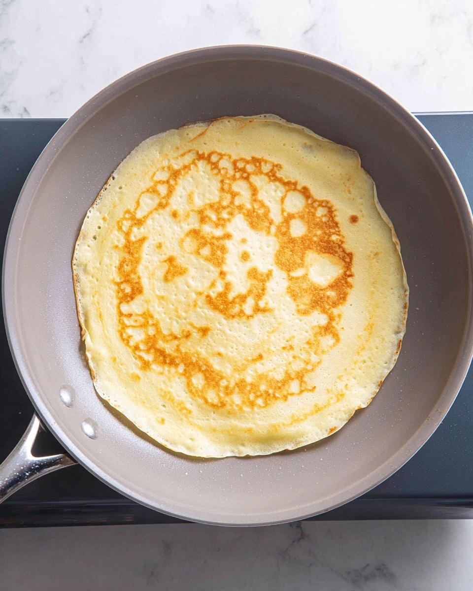 A single thin pancake is cooking in a light gray frying pan with a smooth surface and slight shine, placed on a stovetop with a white marbled texture beneath. The pancake is pale yellow with uneven light brown spots across its surface, showing thin, soft texture with slightly crispy edges curling upwards. The pan handle extends out to the left side. Photo taken with an iphone --ar 4:5 --v 7