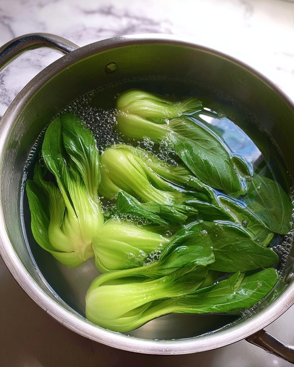 A silver metal pot filled with clear boiling water holding several whole stalks of bright green bok choy with pale green stems and darker green leafy tops visible under the bubbling water; the pot is placed on a surface with a white marbled texture, and light reflects softly off the shiny water and metal edges photo taken with an iphone --ar 4:5 --v 7