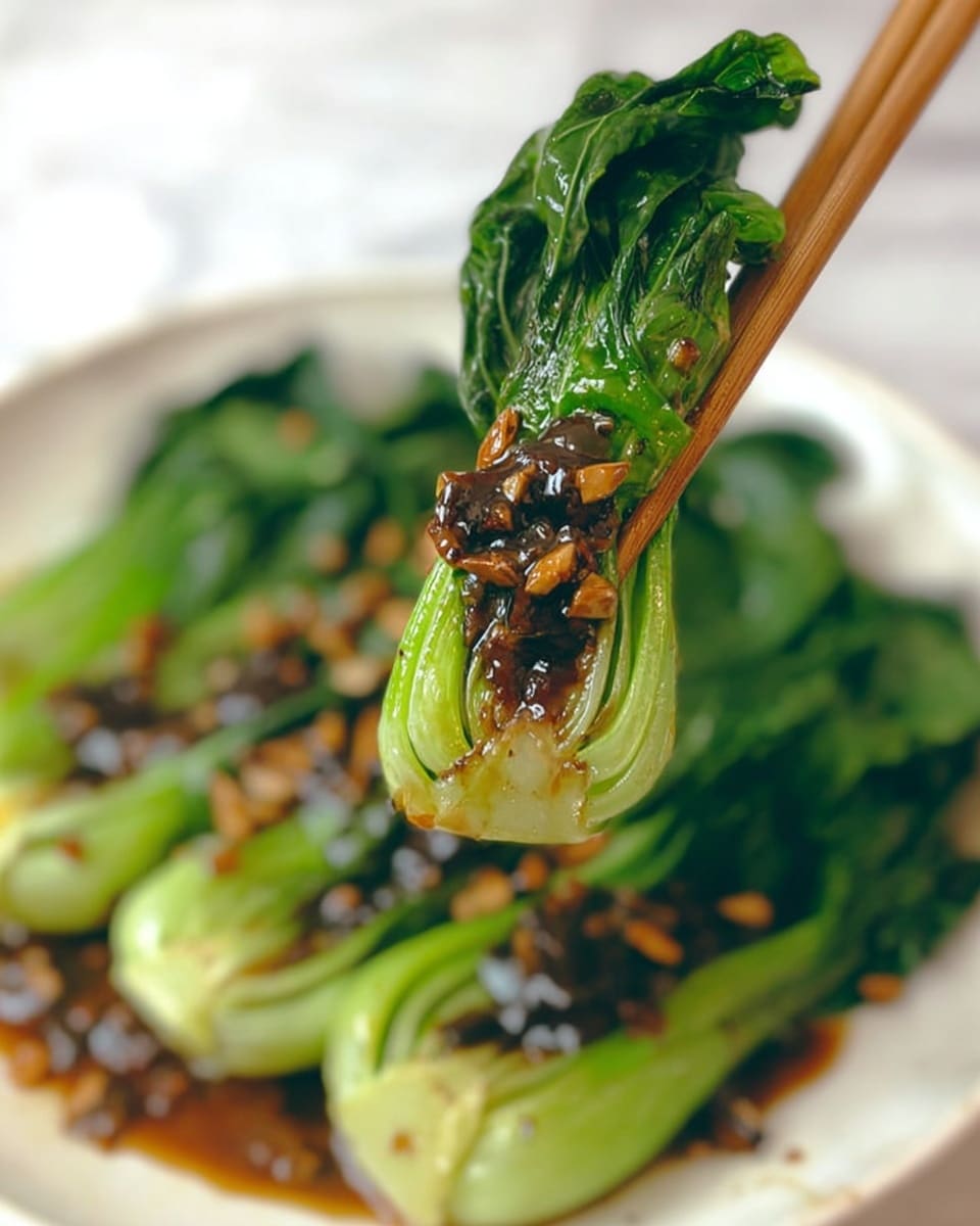 A piece of bright green bok choy with visible leafy layers is held by a pair of wooden chopsticks above a white plate. The bok choy is topped with a glossy, dark brown sauce that has small bits of garlic or nuts on it, adding texture. The plate underneath holds more pieces of bok choy arranged with their green leaves spread out and coated in the same sauce, all placed on a white marbled surface. The focus is on the bok choy in the chopsticks with a soft blurred background. Photo taken with an iphone --ar 4:5 --v 7
