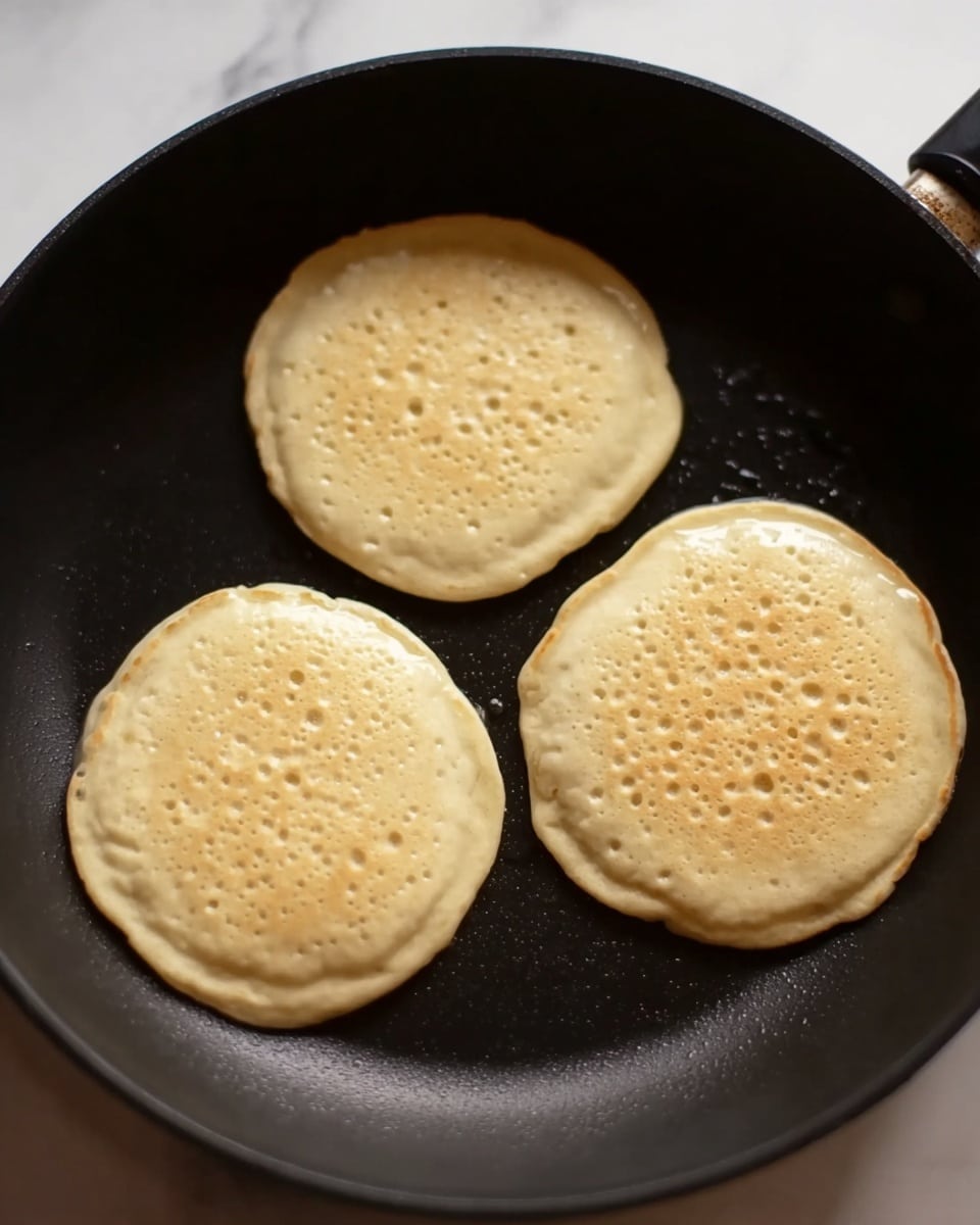 In the image, there are three thick pancake batters cooking inside a black frying pan. Each pancake is light beige with small bubbles and uneven surfaces showing they are still cooking. The pancakes are round but have soft, natural edges, and the pan’s dark texture contrasts with the light batter. The background is a white marbled texture. photo taken with an iphone --ar 4:5 --v 7
