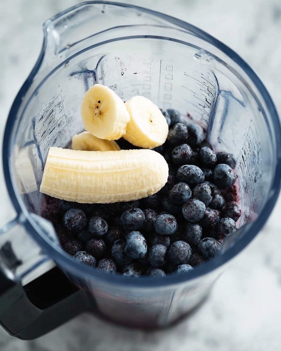 A clear blender jar filled with dark purple-blue round blueberries forming the bottom right layer and two peeled banana halves resting diagonally on top, showing pale yellow, soft texture. The blender is placed on a white marbled surface that gives a clean and fresh look. The background is softly blurred, focusing attention on the fresh fruit inside the jar. Photo taken with an iphone --ar 4:5 --v 7