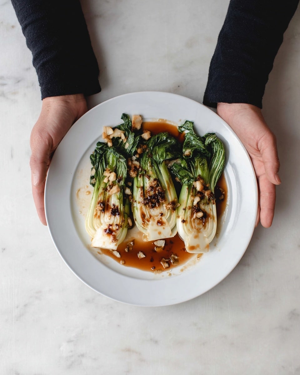 The image shows a white round plate on a white marbled surface with two pieces of cooked bok choy. Each bok choy is cut in half lengthwise, showing a creamy white base with green leafy tops that are slightly wilted and browned from cooking. The bok choy is placed side by side with a drizzle of brown sauce and small bits of cooked garlic scattered on and around the vegetable halves. A woman's hand is placed on each side of the plate, with both hands resting flat on the surface. The person wears a black long-sleeve top. photo taken with an iphone --ar 4:5 --v 7